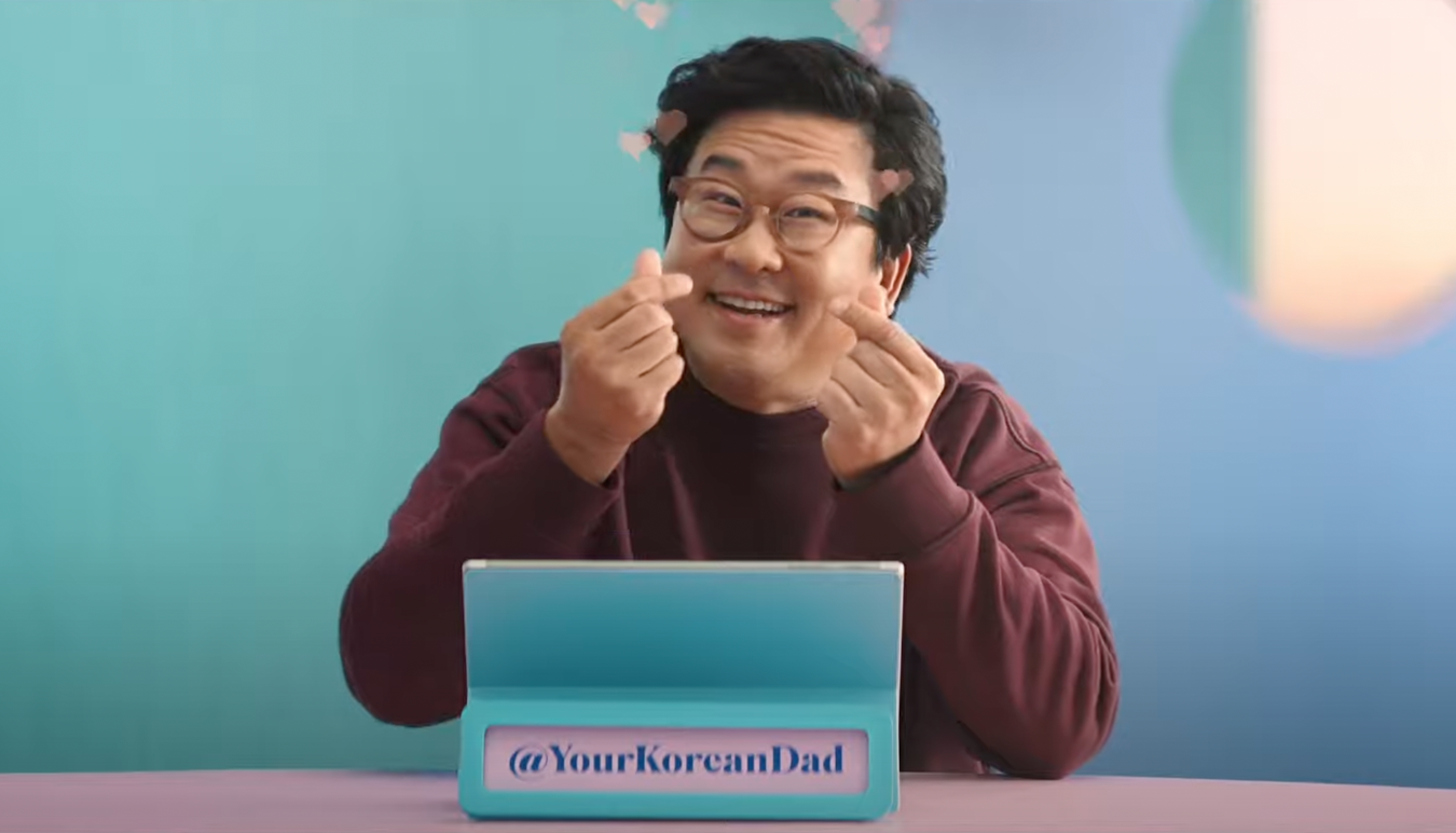 Nick is shown holding up two finger hearts. There's a sign in front of him which reads "Your Korean Dad."